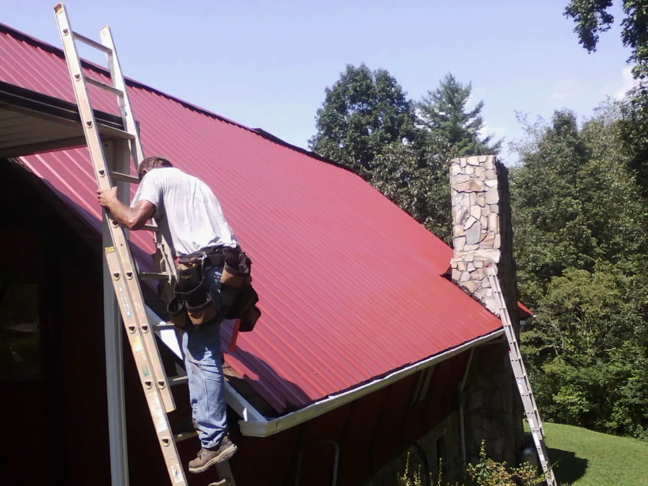 Roofer on a ladder beside a red metal roof and stone chimney — Soffit & Fascia Repair by Roofing Brothers in Valley
